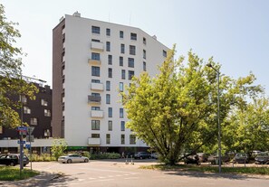Ein mehrstöckiges Gebäude mit flachem Dach, mehreren Balkonen und einem Parkplatz mit Autos.