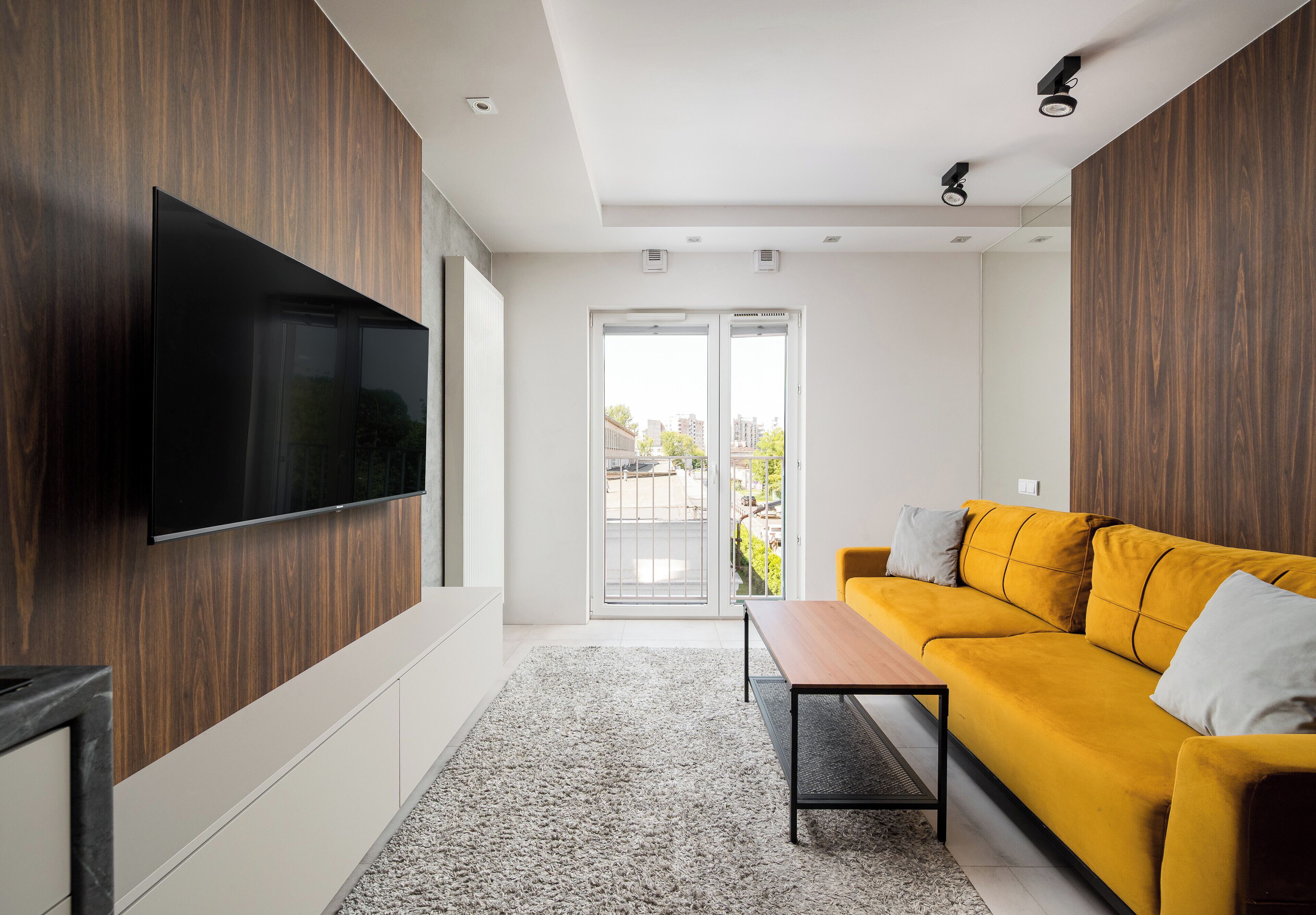 Una sala moderna con televisor de pantalla plana, un sofá amarillo, un muro de acento de madera y un portón corredizo de vidrio que da a un espacio exterior.
