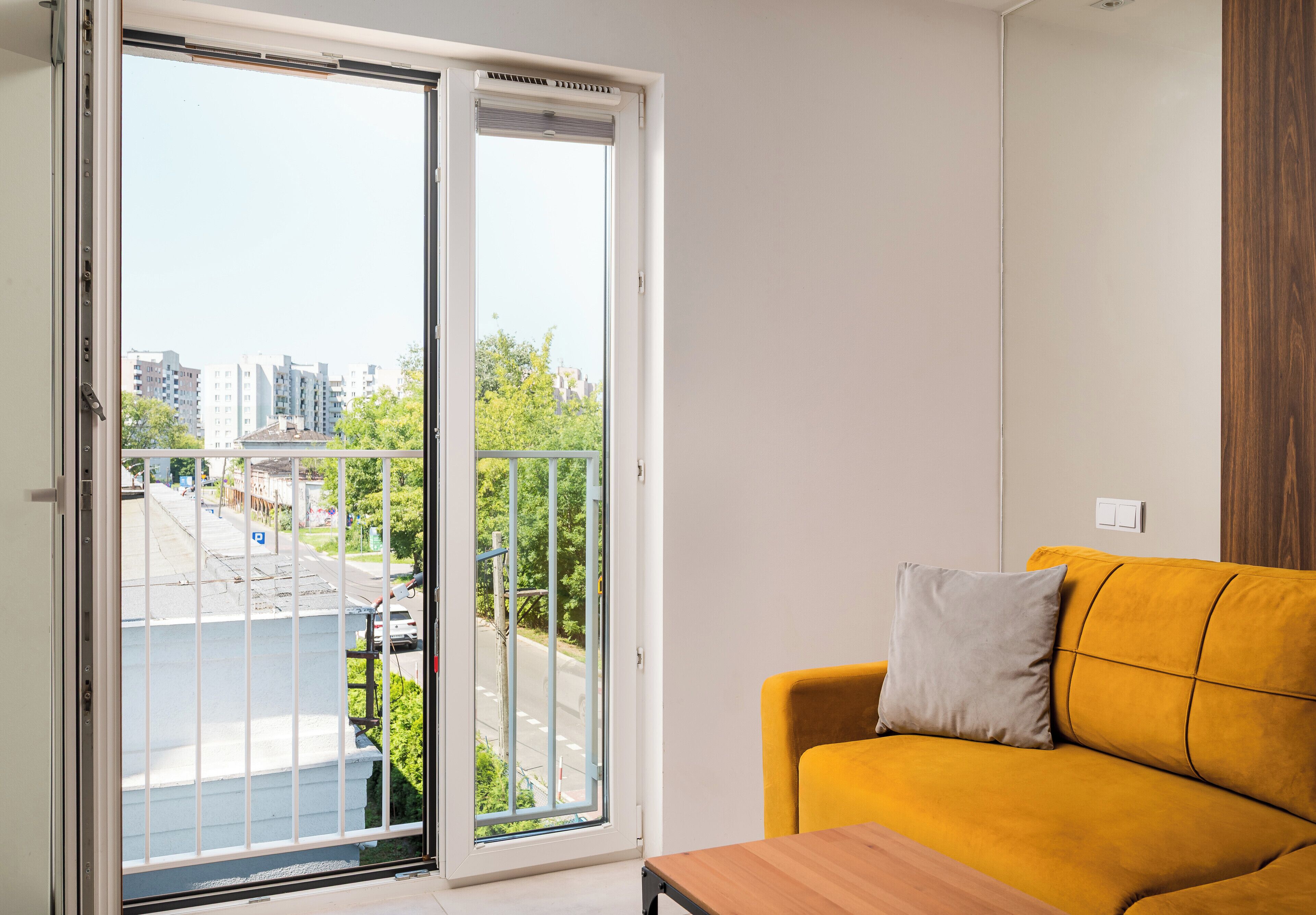 A room with a yellow sofa, a wooden table, and a sliding glass door leading to an outdoor area with a view of buildings and trees.