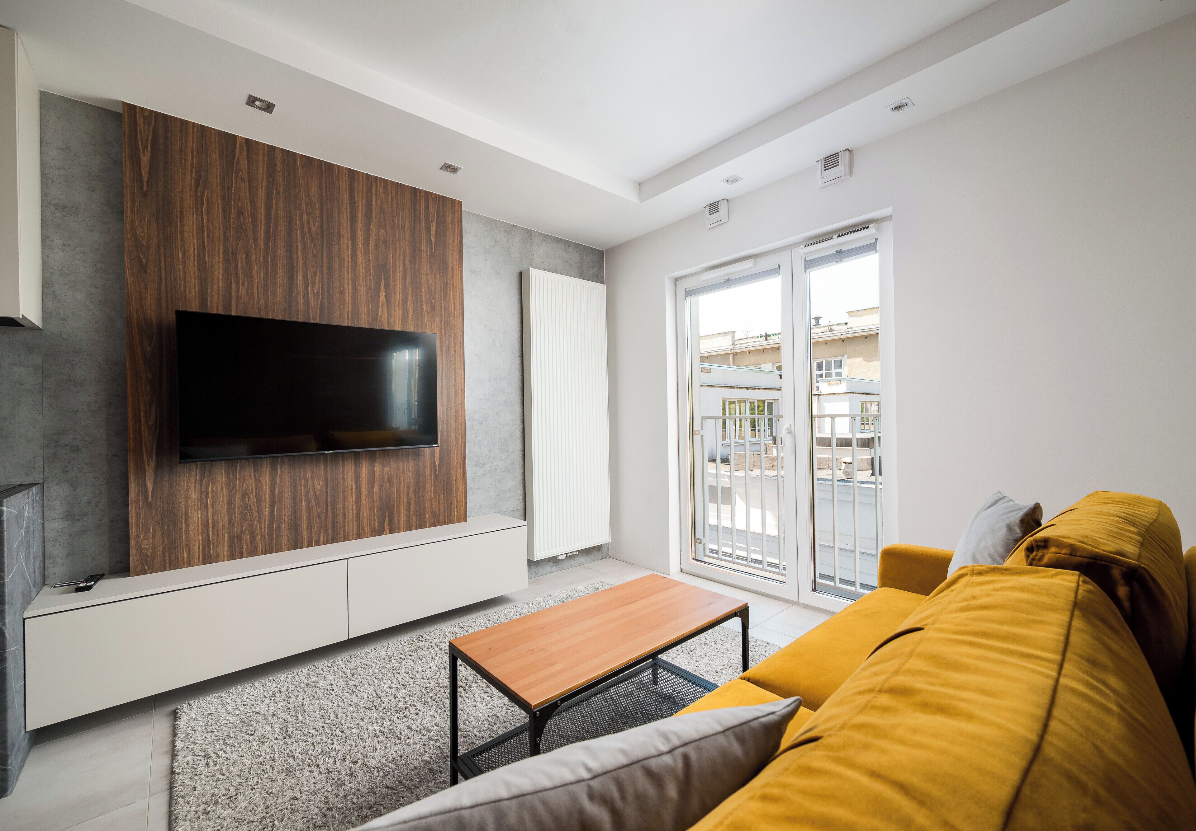 A modern living room with a flat-screen TV mounted on a wooden panel wall, a sofa, a coffee table, and a sliding glass door leading to an outdoor area.