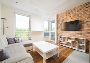 A modern living room with a white sofa, a coffee table, a brick wall, a TV mounted on the wall, and a large window. - Pieskowa Skała 15 Prestige Apartment (Warszawa)