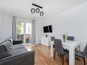 Ein modernes Wohnzimmer mit einer Couch, einem Esstisch mit Stühlen, einem an der Wand montierten Flachbildfernseher und einer Glastür zum Balkon.
