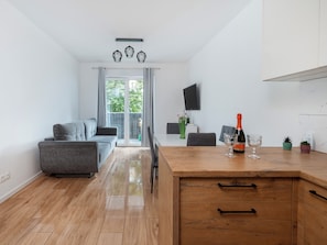Ein modernes Wohnzimmer mit einem grauen Sofa, einem Holzesstisch mit Flasche und Glas sowie einem an der Wand befestigten Fernseher.