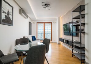 Un salon moderne avec une table en verre, des fauteuils gris, un canapé, un téléviseur posé sur une étagère et une grande fenêtre donnant sur un beau paysage.