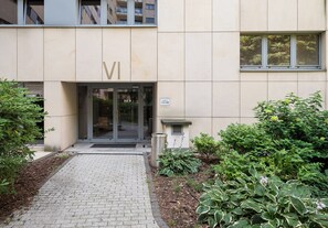 L’entrée d’un immeuble, avec un chemin pavé menant à une porte en verre, entourée de verdure et agrémentée d’une poubelle en métal.