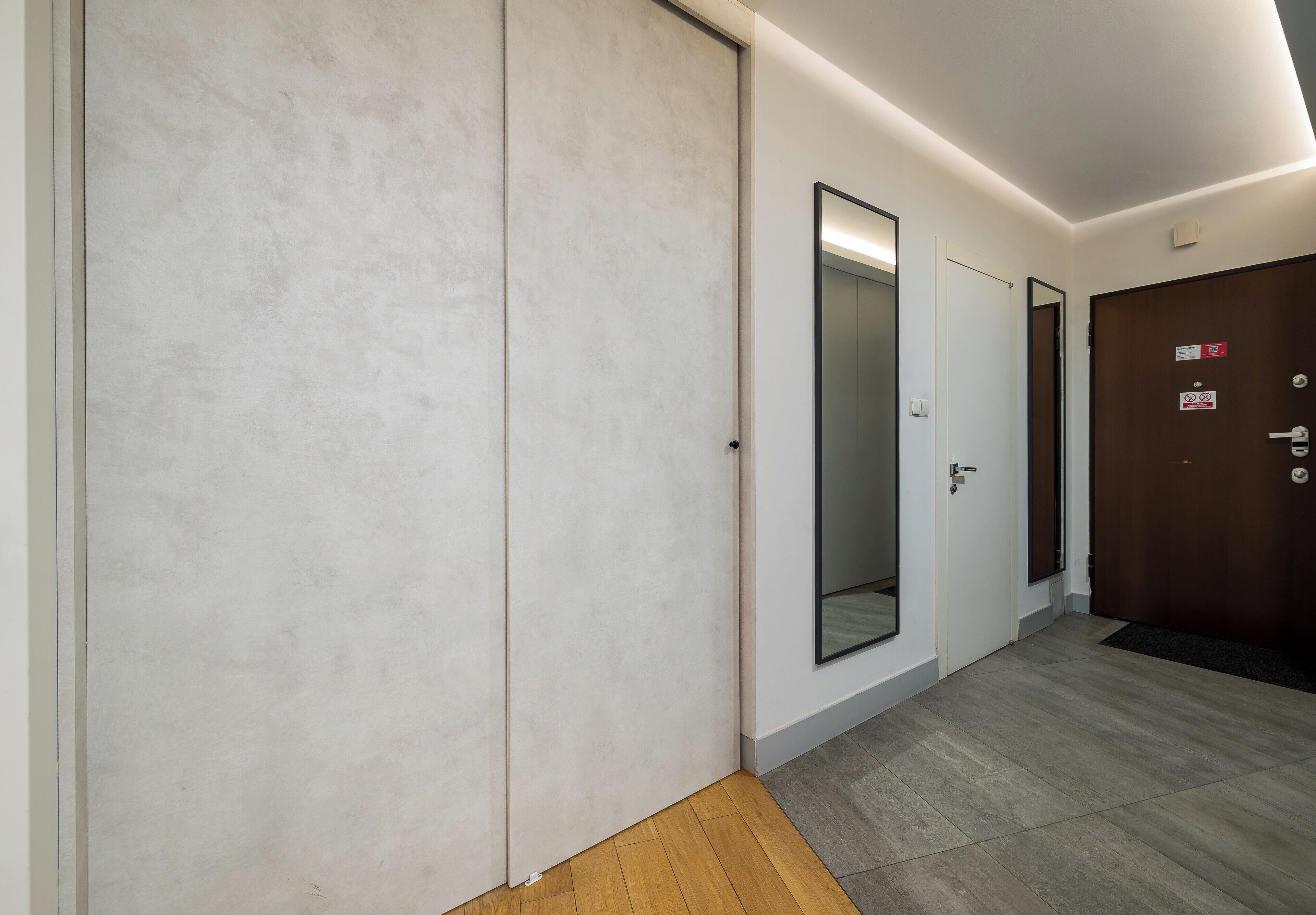 A hallway with a wooden floor, a white wall, and a door with a mirror.