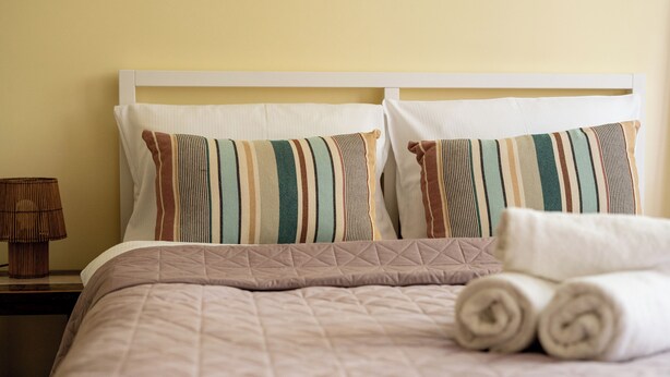 Una cama bien hecha con almohadas a rayas y una toalla enrollada, una lámpara de noche y una pared lisa.