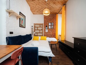 Une pièce comprenant un lit, un bureau, une chaise, une commode, un luminaire suspendu et un mur en briques orné de tableaux.