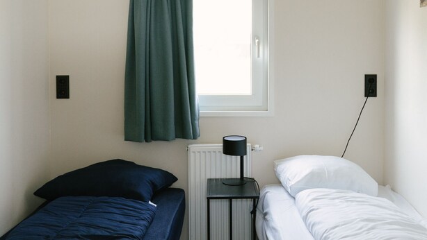 Une petite chambre d’hôtel, composée d’une seule pièce, comprenant un lit, une table de chevet, une lampe et une fenêtre avec des rideaux.