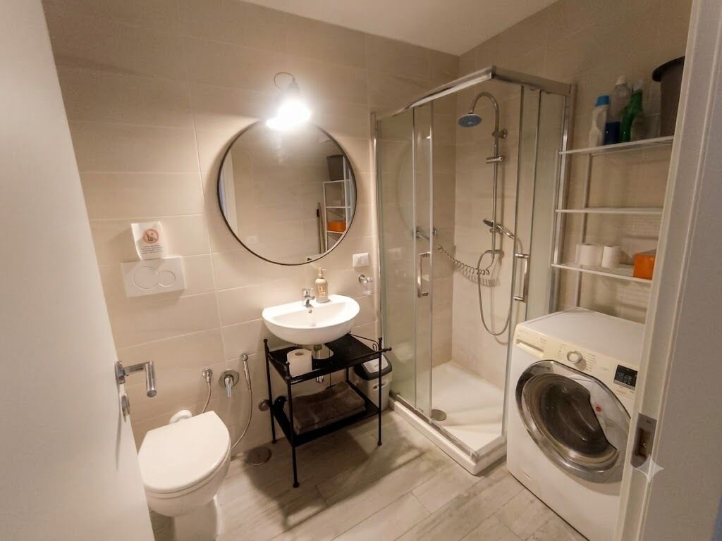 A bathroom with a toilet, sink, mirror, shower, and a washing machine.