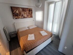Ein Schlafzimmer mit einem Bett, Kissen, einem Nachttisch, einem Wandbild, einem Fenster mit Jalousien und einer Tür.