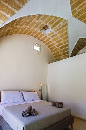 Une chambre à coucher avec un plafond orné d’un motif en briques, un lit recouvert de draps blancs et une petite fenêtre.