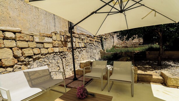 Un espacio de estar al aire libre con un muro de piedra de fondo, una mesa de madera y un ramo de flores, además de un paraguas grande que da sombra.