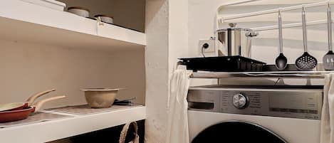 A compact laundry room with a washing machine, shelves with kitchenware, and a small trash bin.