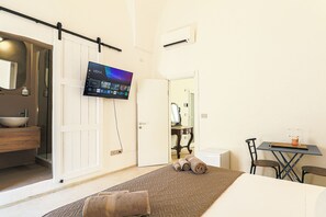 Ein Hotelzimmer mit einem Bett, einem Schreibtisch, einem Sessel, einem an der Wand montierten Fernseher sowie einem Badezimmer mit Waschbecken und Spiegel.
