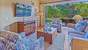 A living room with a flat-screen TV, wooden furniture, a glass door leading to a garden, and a view of greenery. - Maui Resort Rentals: Two Luxury Ground Floor Suites with Expansive Lawns Near Beach, Pools and BBQs! (Lahaina)