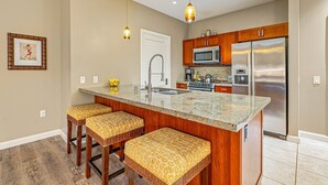 A kitchen with a granite countertop, stainless steel appliances, wooden cabinets, and a bar stool seating area. - Maui Resort Rentals: Two Luxury Ground Floor Suites with Expansive Lawns Near Beach, Pools and BBQs! (Lahaina)