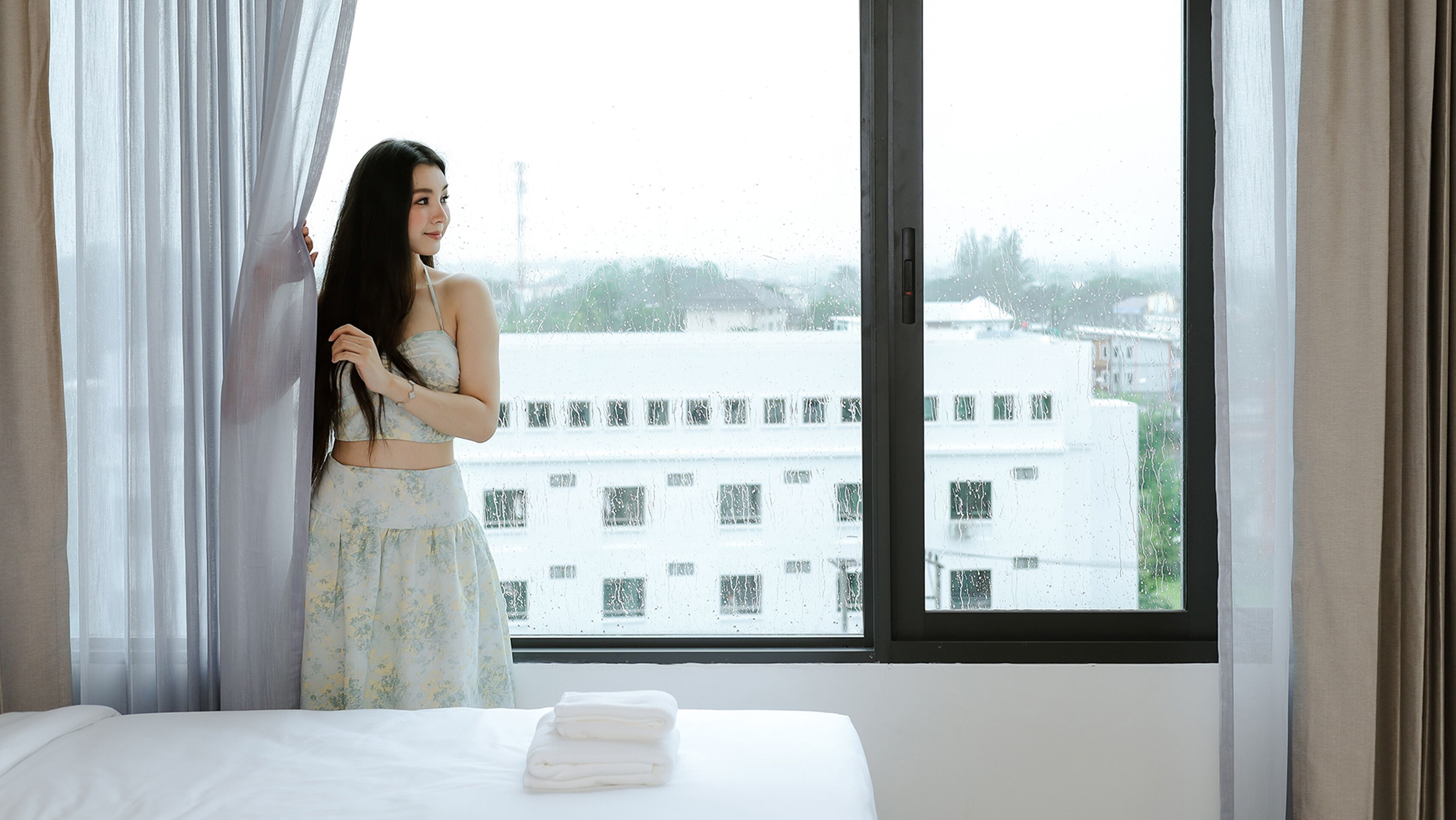 A room with a bed, a window with curtains, and a view of buildings outside.