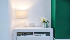 Valkoinen yöpöytä, jossa on lamppu, maljakko kukkineen ja laatikko, jonka päälle on kirjoitettu sanat »PARIS» ja »LONDON».