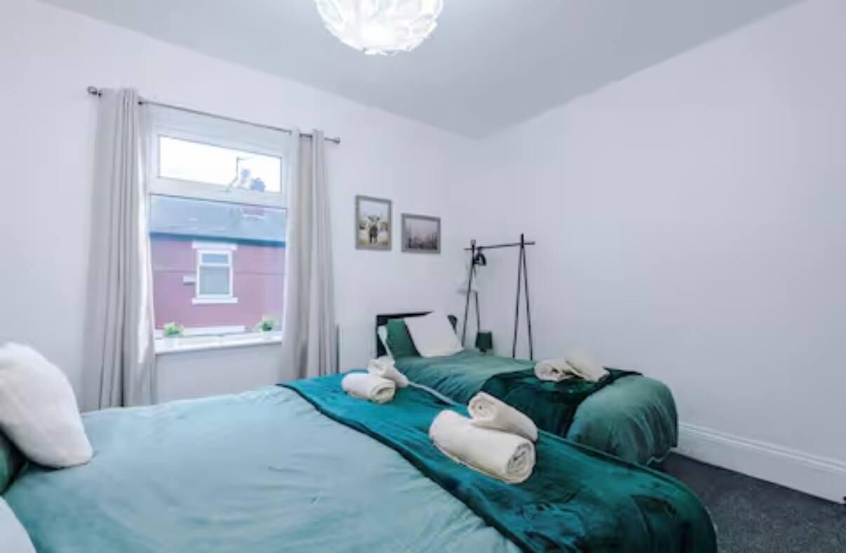 Un dormitorio con dos camas, una ventana con cortinas, una lámpara fijada a la pared y un cuadro pequeño.