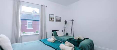 Un dormitorio con dos camas, una ventana con cortinas, una lámpara fijada a la pared y un cuadro pequeño.