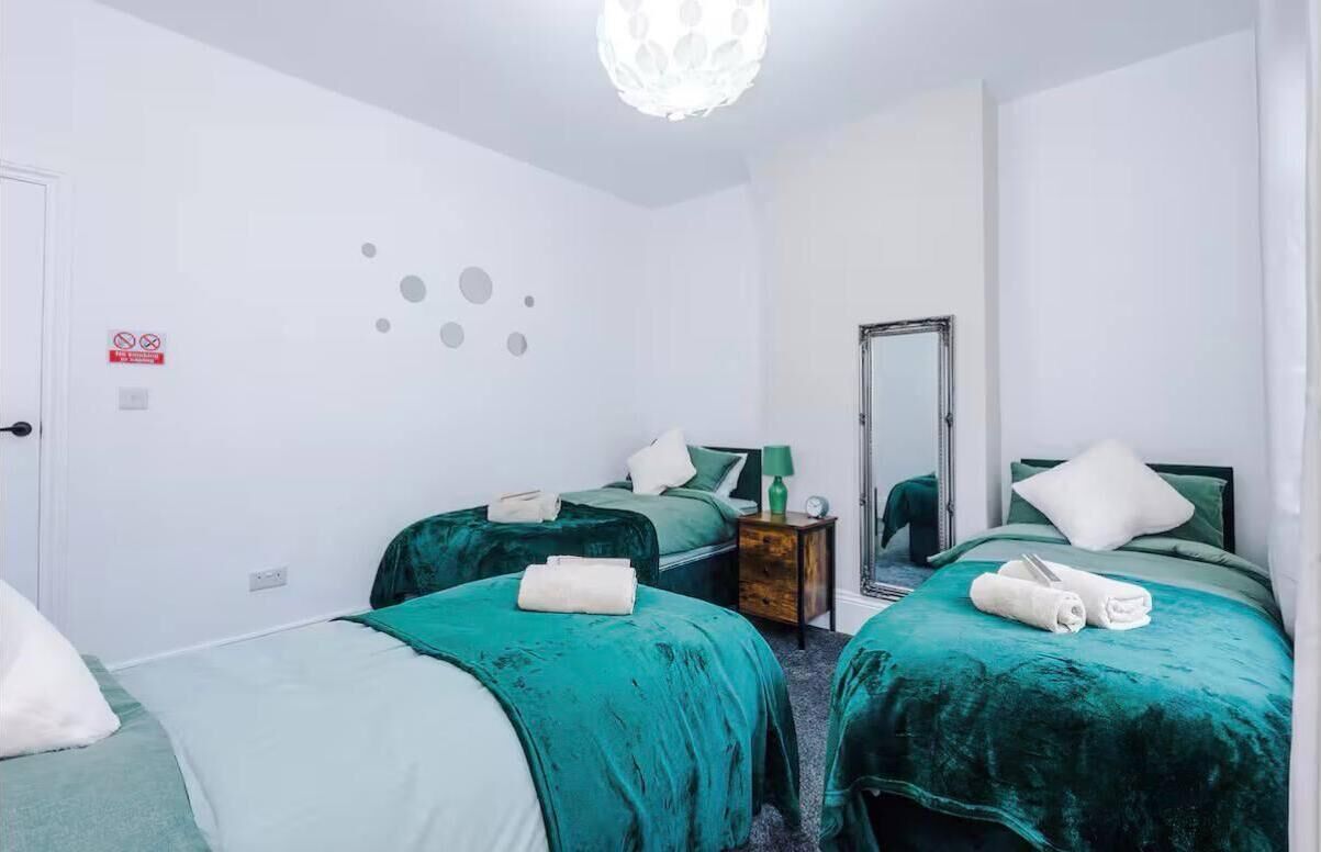 A hotel room with two beds, each with a green bedspread and white pillows. There is a small wooden nightstand with a green lamp and a clock. The room has white walls and a white door.