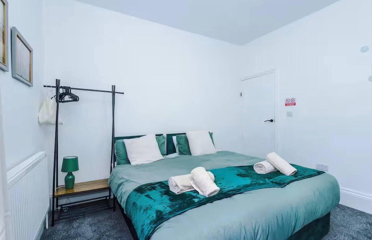 Een enkel bed met een groene sprei en kussens, een nachtkastje met een lamp, een radiator en een deur met een rookverbodsbord.