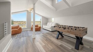 A modern room with a foosball table, a white sofa, and a view of the desert through large windows. - Sanibel Beach Retreat + Bonus Bungalow! Sleeps 17 • Private Pool (Sanibel)