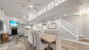 A spacious dining area with a long table and wicker chairs, a television mounted on the wall, and a staircase with white railings. - Sanibel Beach Retreat + Bonus Bungalow! Sleeps 17 • Private Pool (Sanibel)