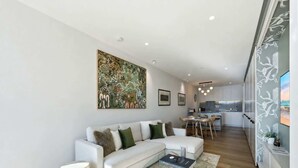 Ein modernes Wohnzimmer mit einem weißen Sofa, einem Glastisch und einem Essbereich, im Hintergrund ist eine Küche zu sehen.