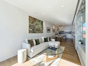 A modern living room with a white sofa, a glass coffee table, and a dining area with a kitchen in the background. - Waterfront 1-Bed Apartment in Kingston (Kingston)