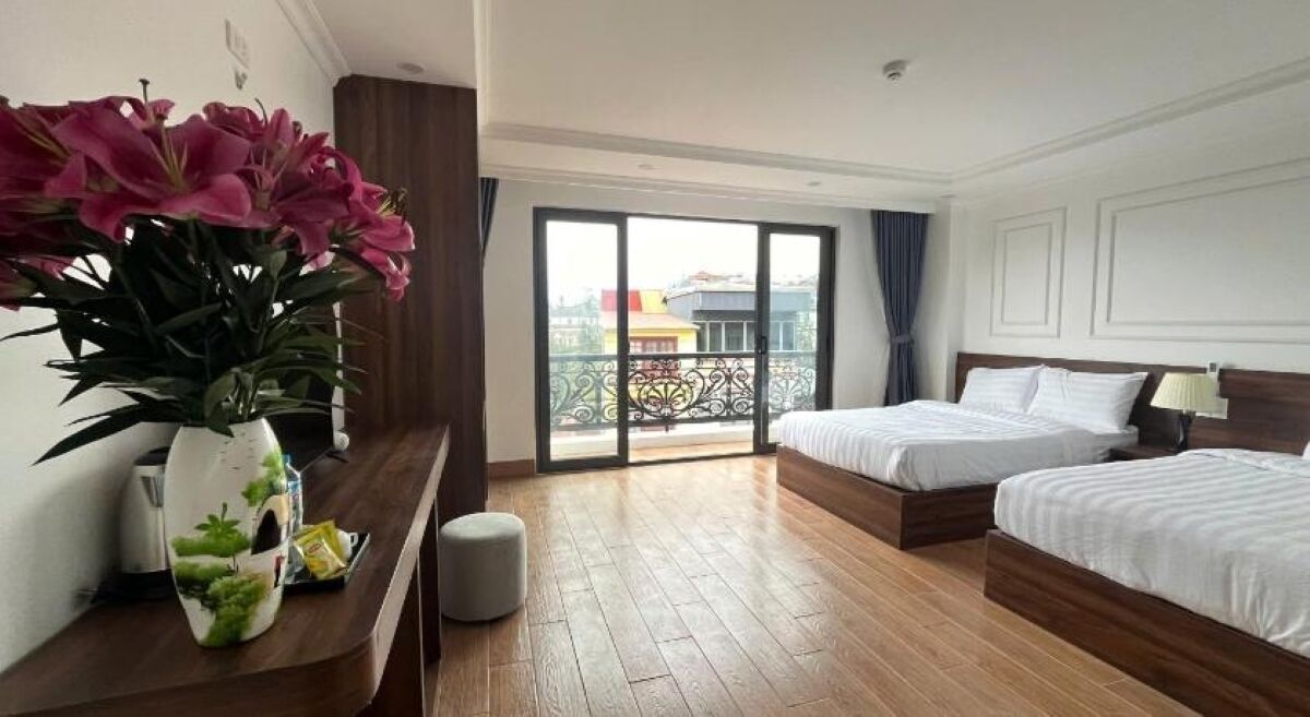 A hotel room with two beds, a wooden floor, a balcony with a view, and a vase with flowers on the nightstand.