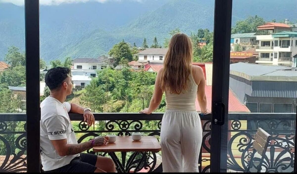 Un balcón con vistas a un paisaje montañoso, una mesa con una taza y un platillo, y una persona sentada en el balcón.