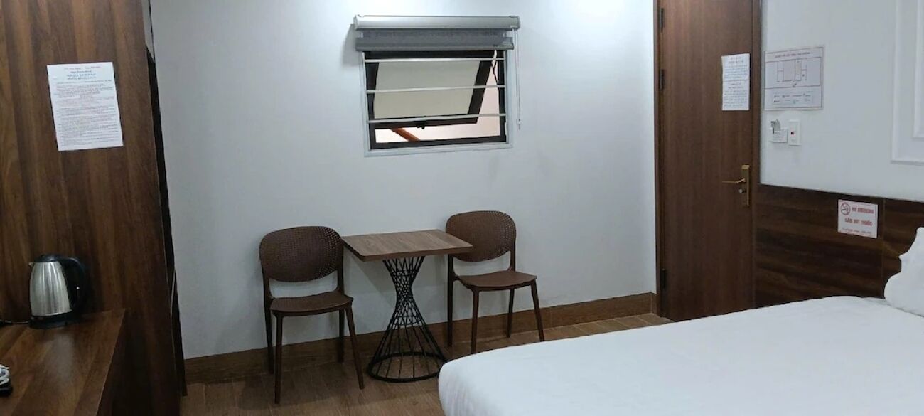Une chambre d’hôtel avec un lit, une petite table et deux chaises, une fenêtre avec un store et une bouilloire posée sur le sol.