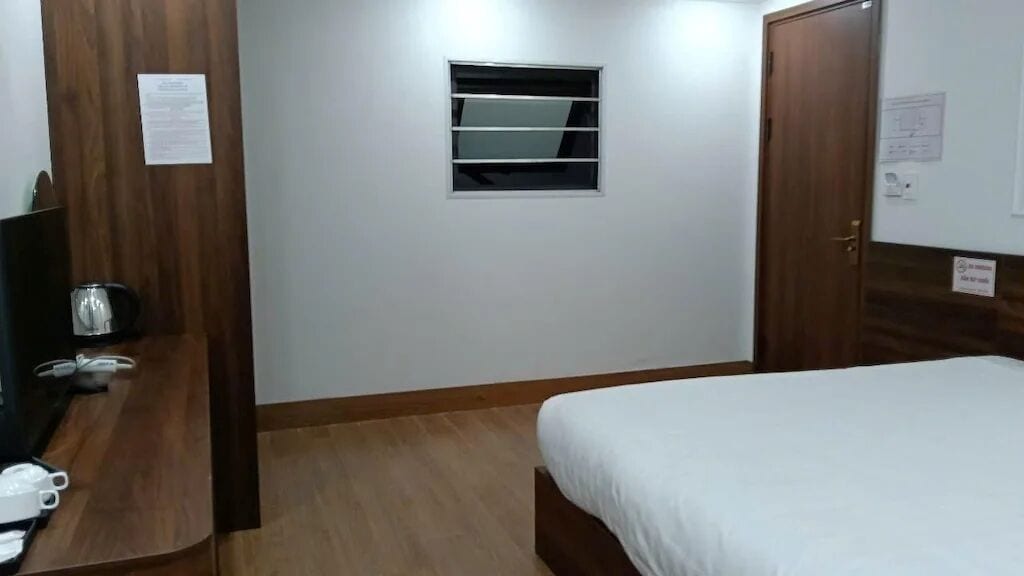 A hotel room with a bed, a small window, a wooden door, and a desk with a kettle.