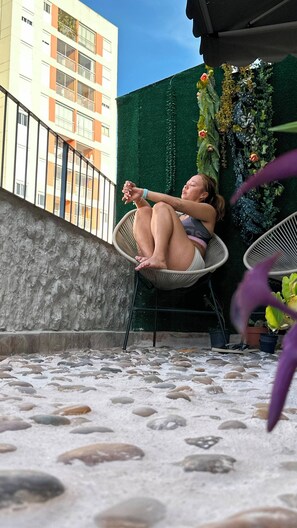 A person sitting on a wicker chair on a balcony with a view of a building and a green wall with plants. - Your apartment in São Paulo | Balcony, subway and the best of downtown (São Paulo)