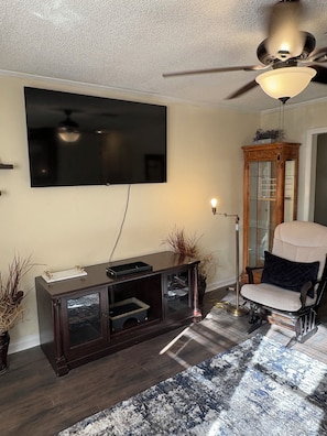 Una sala de estar con un televisor de pantalla plana montado en la pared, un mueble de entretenimiento de madera oscura, un sillón blanco, un tapete estampado y un ventilador de techo.