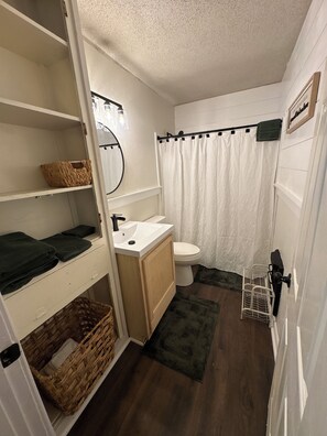 Un baño pequeño con lavamanos, inodoro y cortina de ducha blancos. Hay repisas con cestas y toallas dobladas.