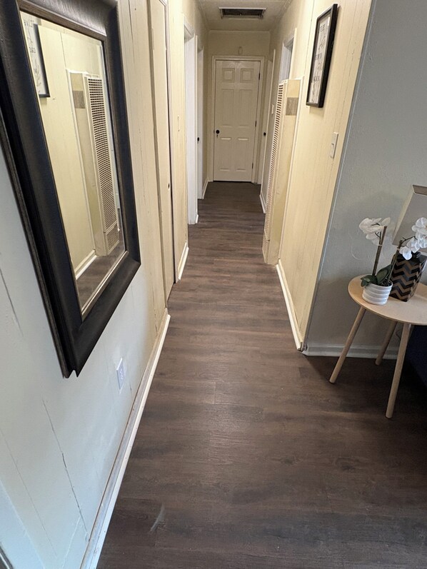 A narrow hallway with a wooden floor, a door at the end, and a small table with a vase on the right side.
