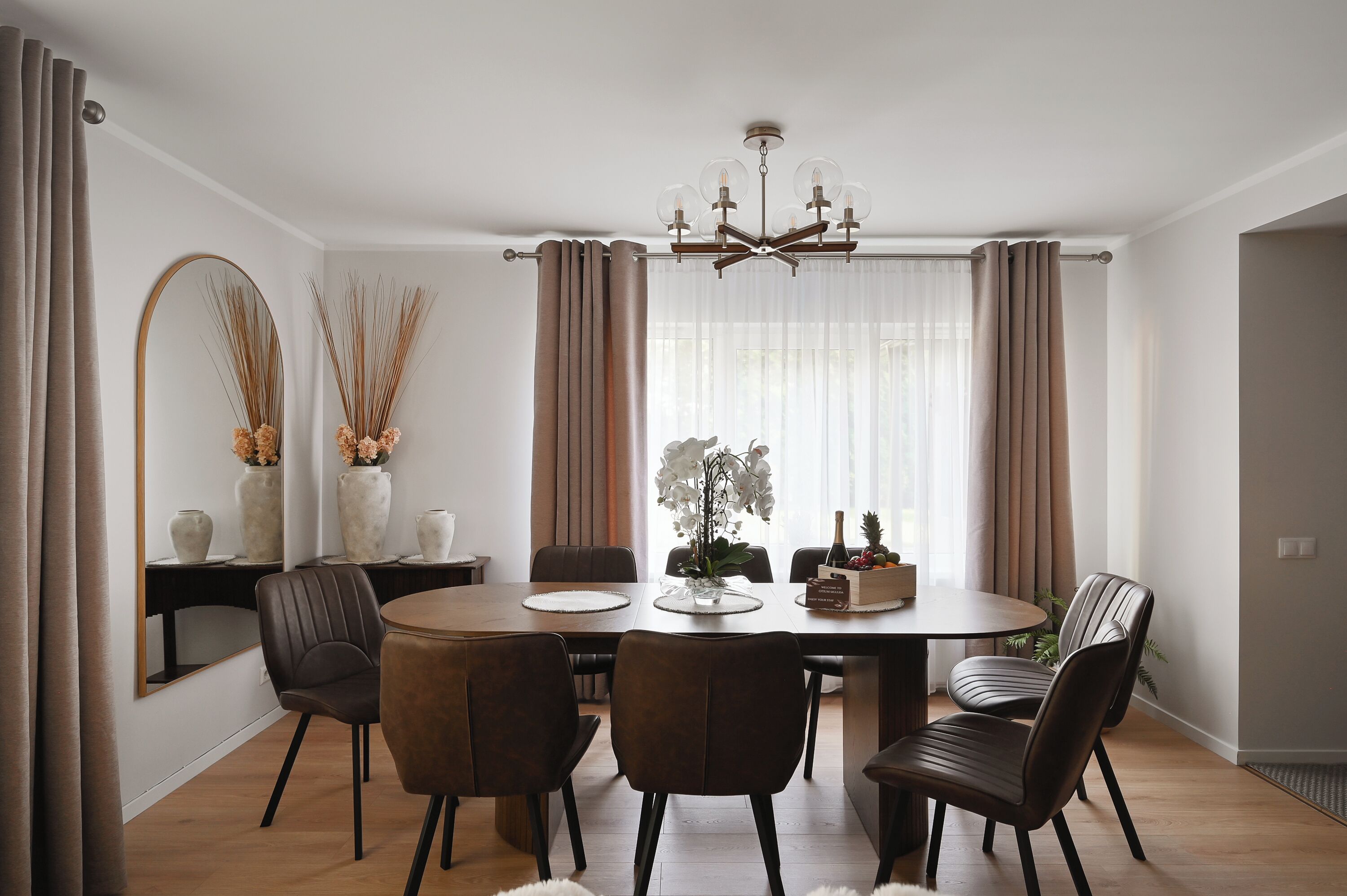 Un coin repas comprenant une table ronde, six chaises, un lustre et une vue sur l’extérieur grâce à de grandes fenêtres.