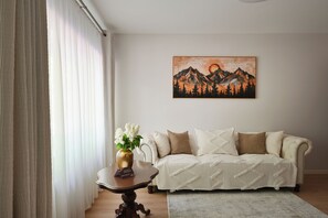 Un salon avec un canapé, une table ornée d’un vase de fleurs et un tableau représentant des montagnes accroché au mur.