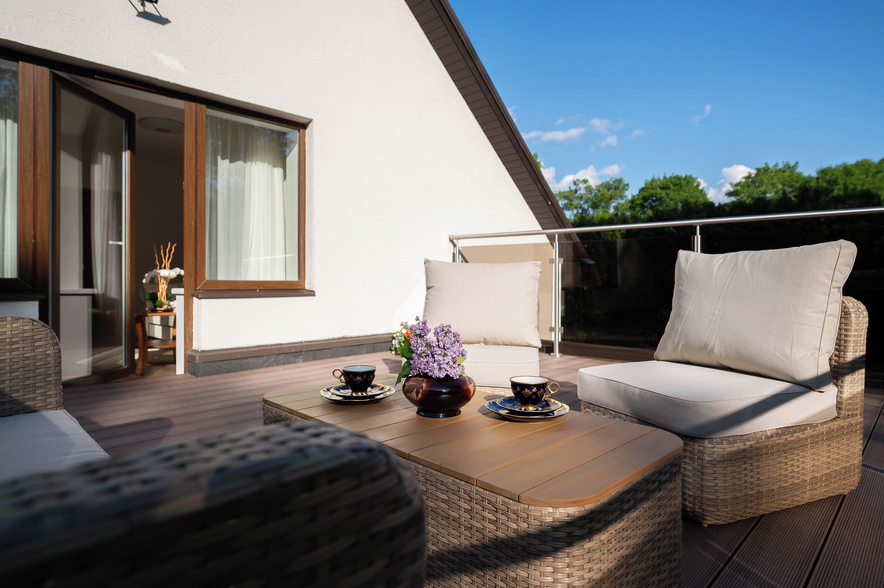 A modern outdoor seating area with wicker furniture, a table with cups and a vase of flowers, and a view of the sky and trees.