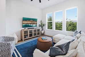 A living room with a large flat-screen TV, a wooden cabinet, a round ottoman, and a sofa with multiple cushions. - Luxury 3BR Waterfront w/ Private Rooftop Skyline Views (Jacksonville)