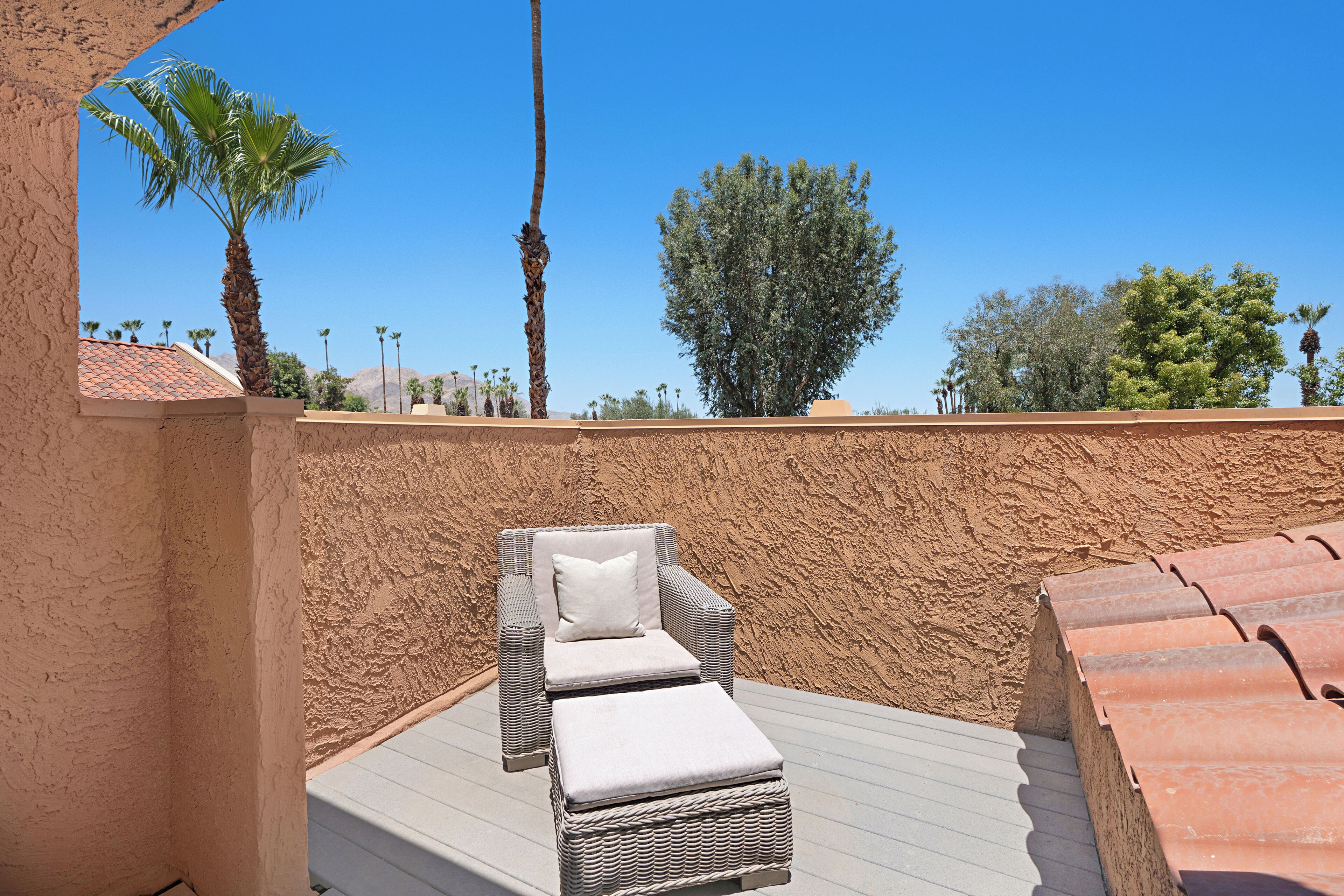 A sunlit outdoor seating area with a wicker lounge chair and ottoman, a textured wall, and a clear blue sky with trees in the background.