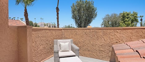 A sunlit outdoor seating area with a wicker lounge chair and ottoman, a textured wall, and a clear blue sky with trees in the background.