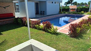 A swimming pool with a tiled deck, surrounded by grass and red-leaved shrubs, with a building in the background.