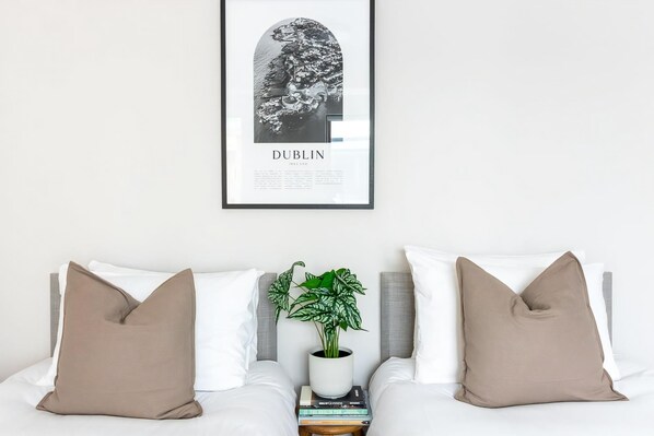 Un cuadro enmarco con una imagen de Dublín en una pared, una cama con ropa blanca y dos almohadas color beige, una planta pequeña en una maceta blanca, y una pila de libros.