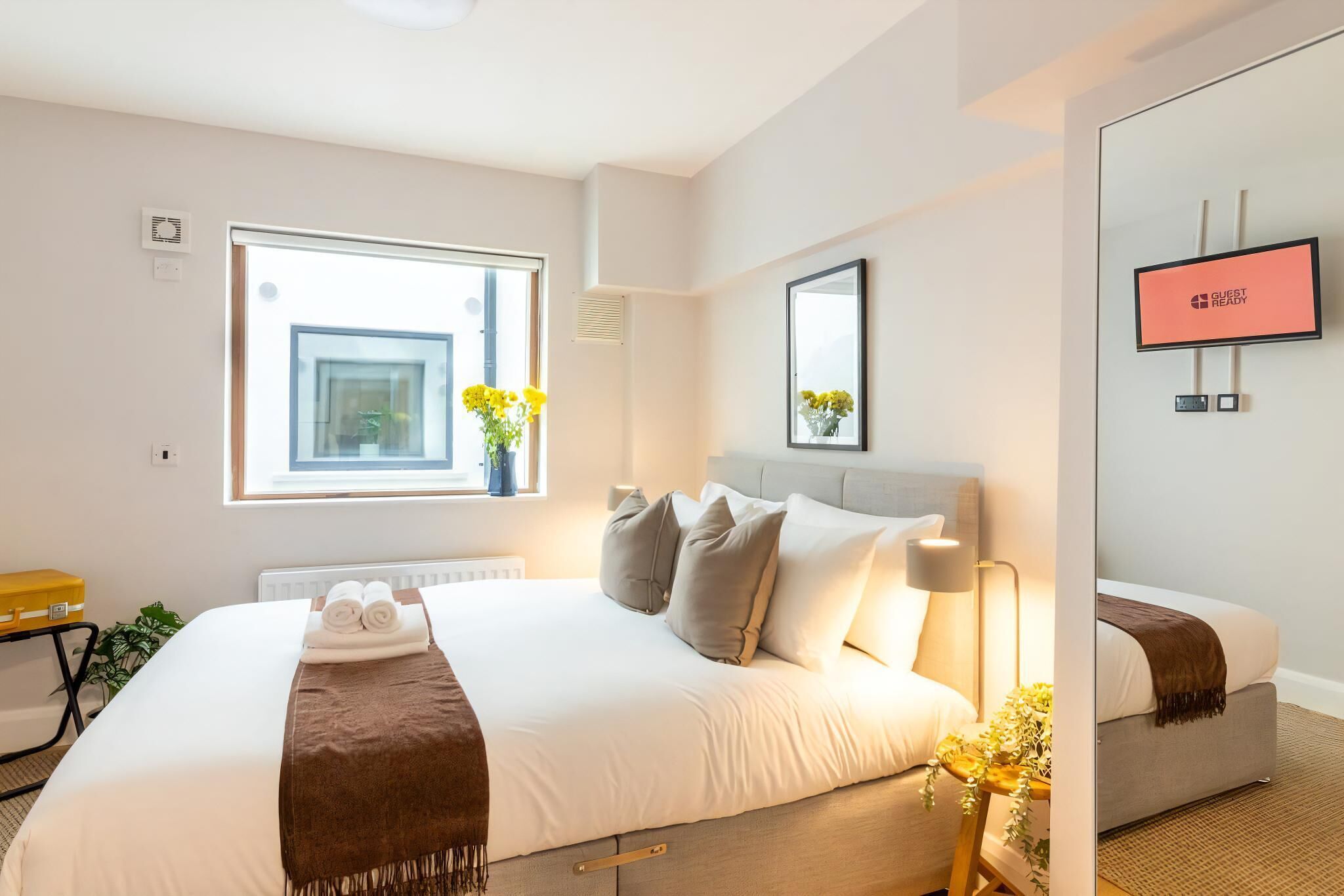 Ett hotellrum med en stor säng, ett nattduksbord med en lampa, ett fönster med blommor och en skylt med texten ”KLART FÖR GÄSTER”.