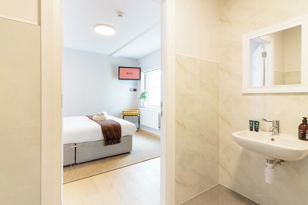 Habitació d'hotel amb un llit, una taula de nit, televisió penjada a la paret i un bany amb un lavabo i mirall.