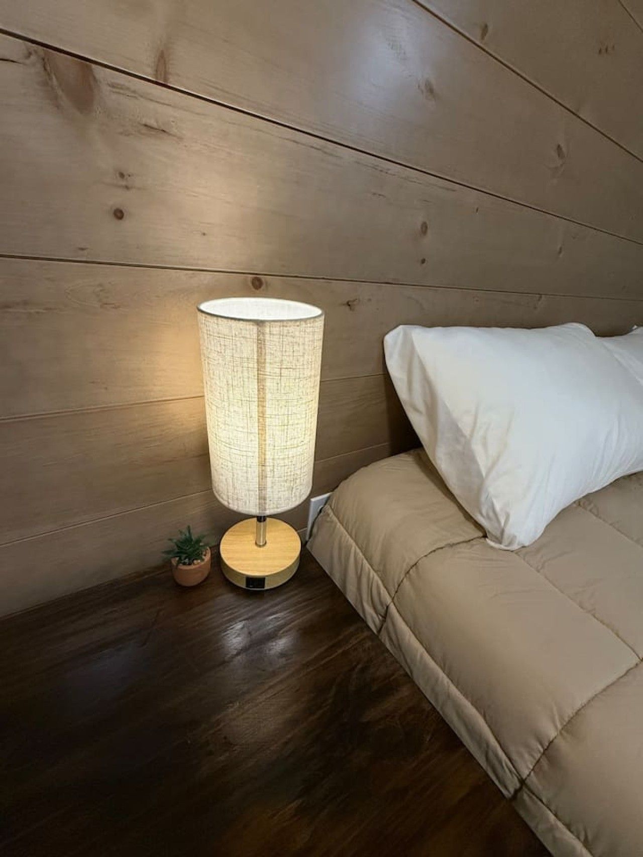 Ein Nachttisch mit Lampe, einer kleinen Pflanze und einem Bett mit weißem Kissen.
